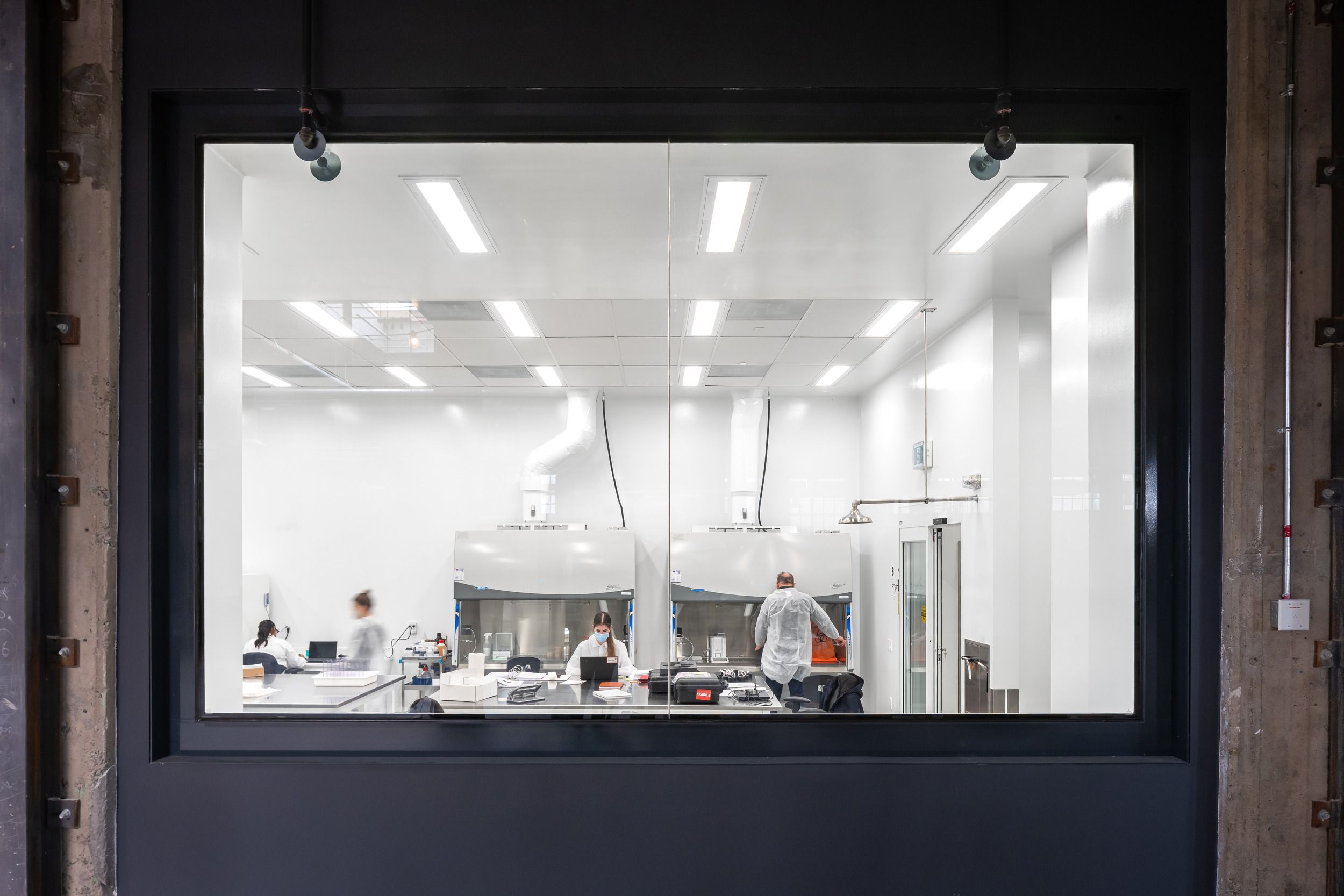 View through observation window into modern scientific laboratory with fume hoods and research equipment.