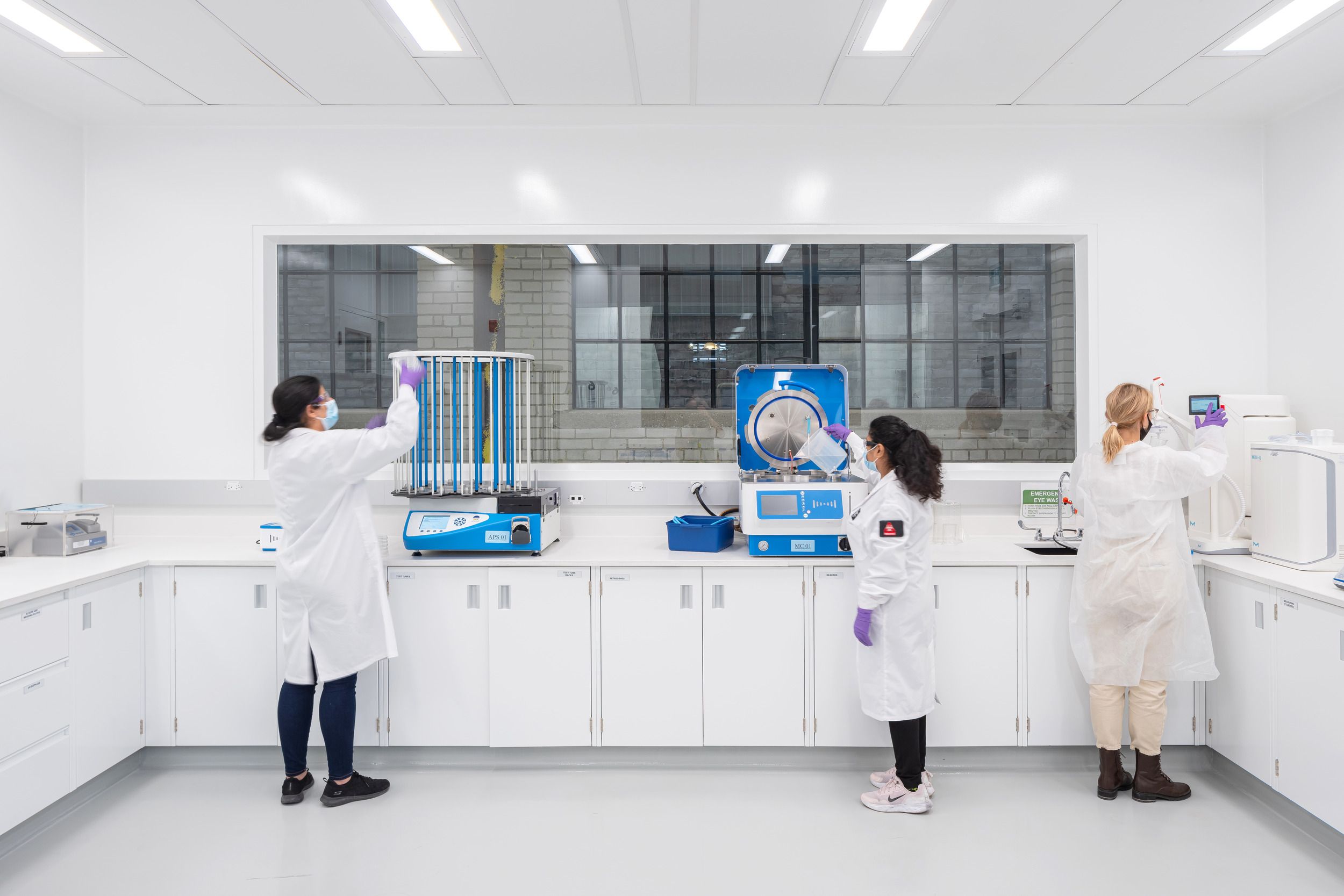Laboratory technicians working with specialized testing equipment in a modern, clean laboratory setting with white counters and cabinets.