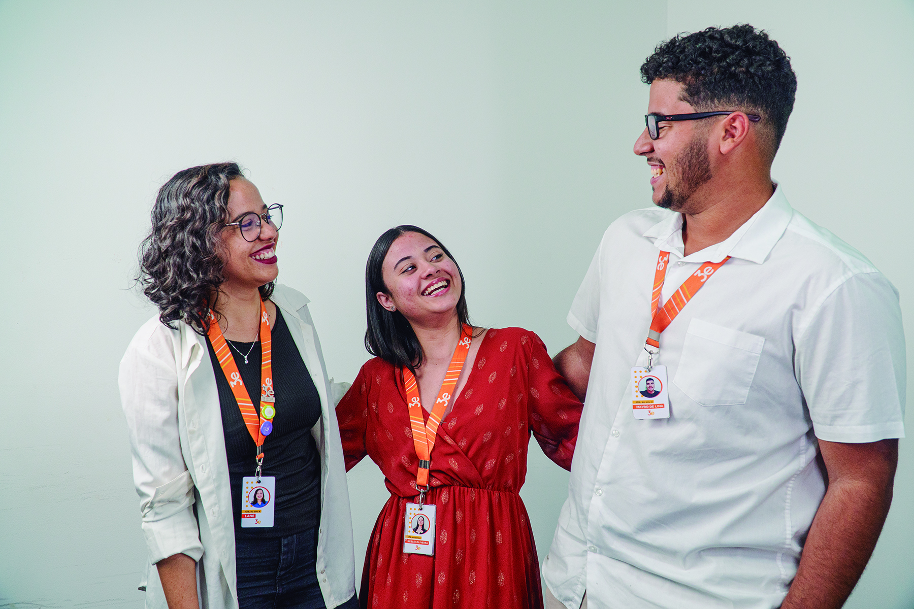 Três colaboradores sorrindo e conversando, usando crachás laranja, em frente a uma parede branca.