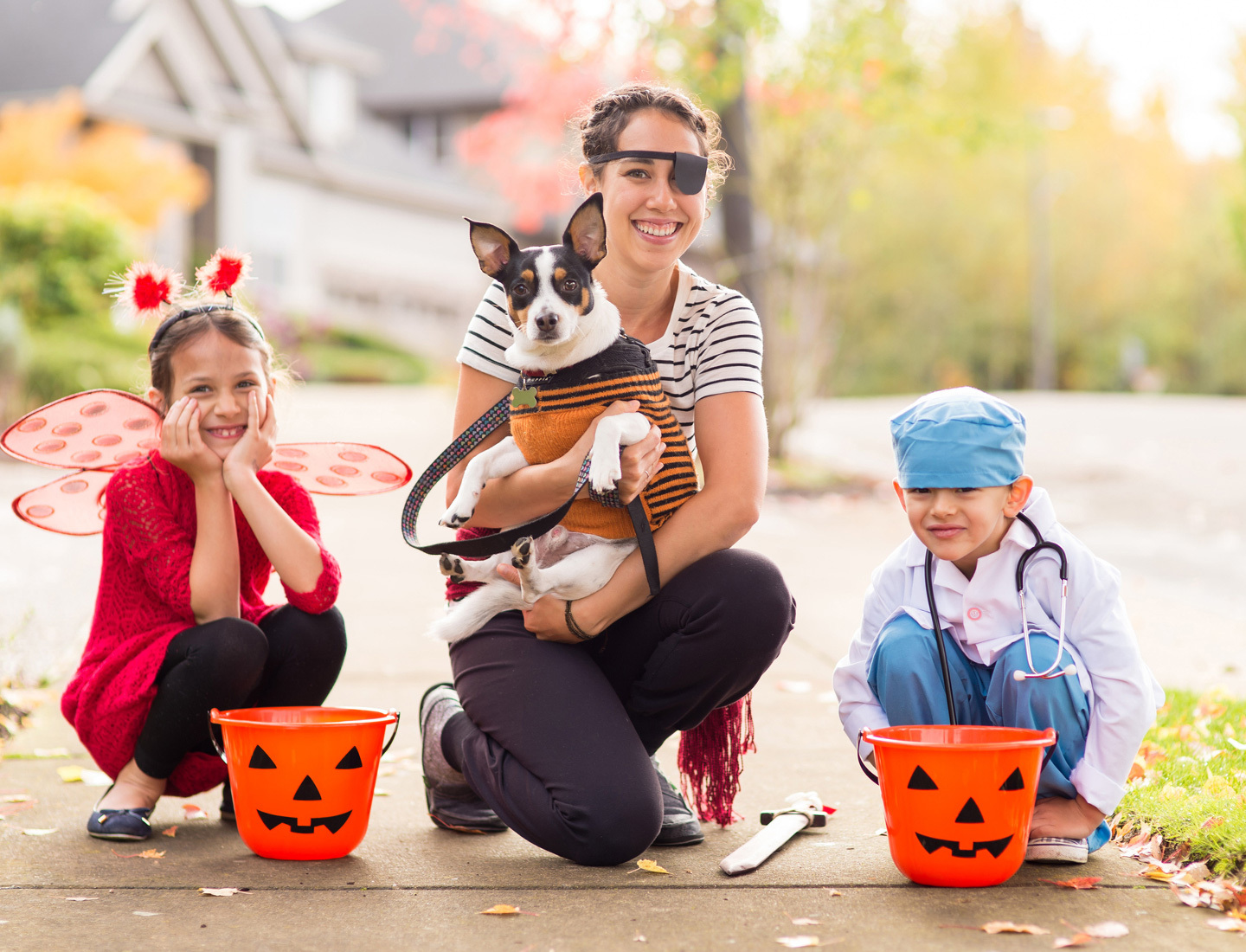 A mother and her two children are in Halloween costumes posing on the sidewalk with their jack-o-lantern candy buckets. The girl is a red insect, and the boy is a doctor in OR scrubs. The mother is a pirate, and she is holding onto their dog.
