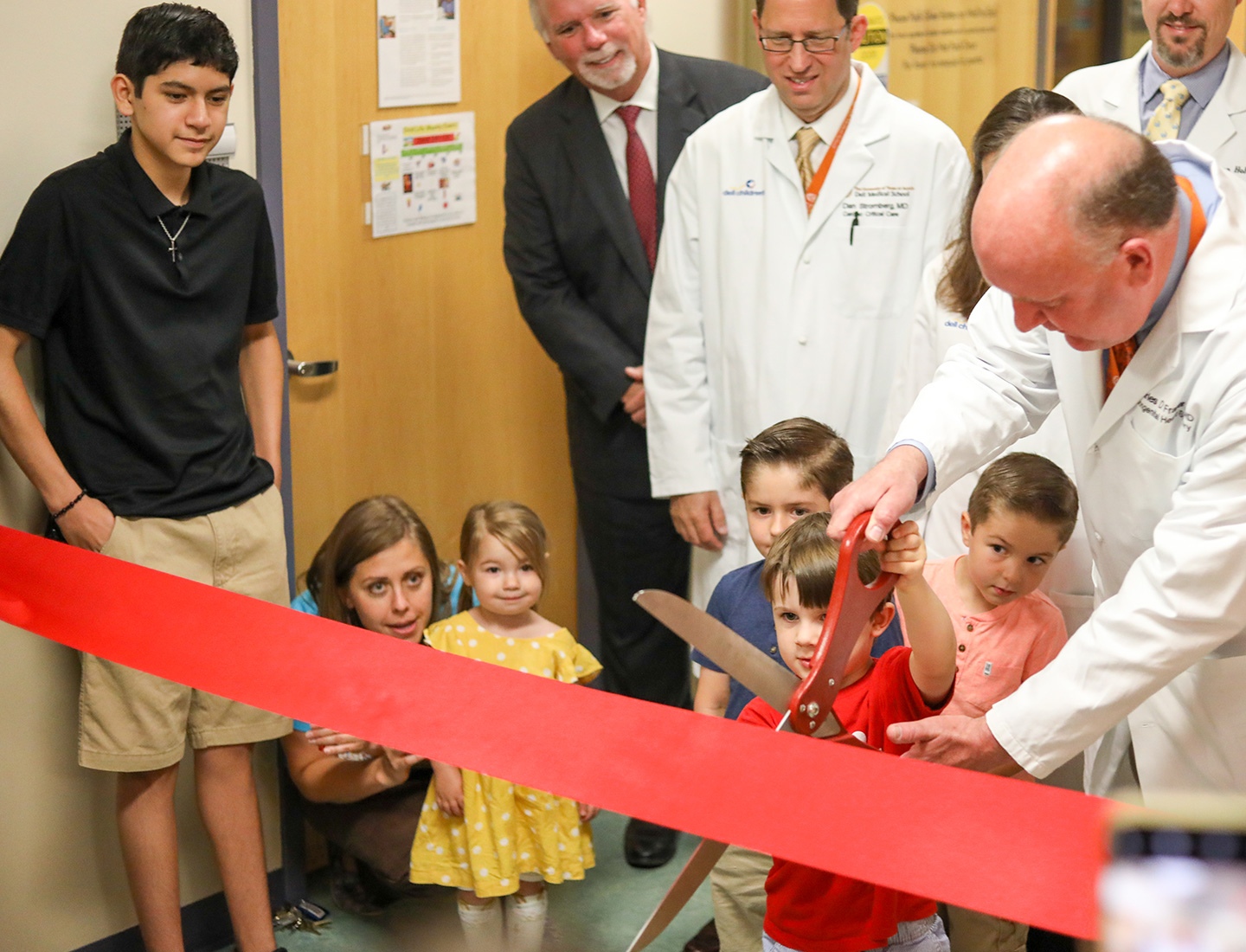 Dr. Charles Fraser is joined by a group of children and other clinicians as he uses an oversized pair of scissors to cut a wide red ribbon that spans the width of a hallway.