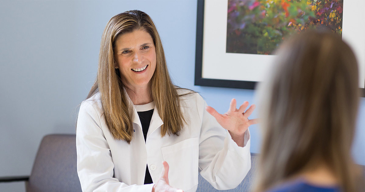 A physician wearing a white coat smiles as she gestures at a patient with her hands. They are seated across from one another in a consultation room with blue walls and natural light filtering through.