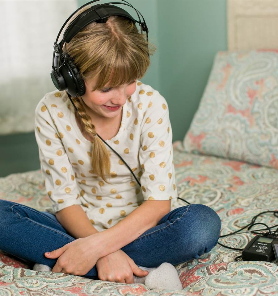 A girl sits cross-legged on her bed, listening on headphones connected to a Waves amplifier