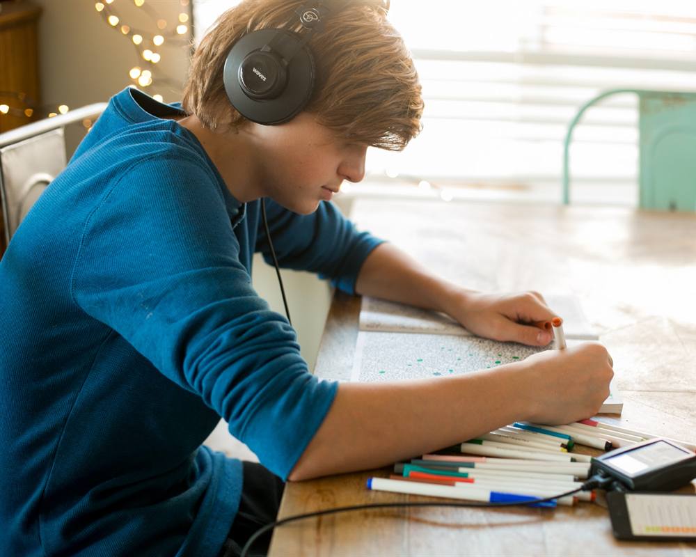 A teenage boy wearing Waves headphones works at a table, with his phone and Waves amplifier beside him