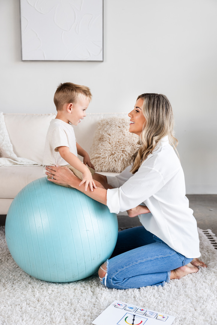 Rachel Harrington kneels on a rug and steadies her young son as he sits on a large blue therapy ball, the two of them smiling at each other.